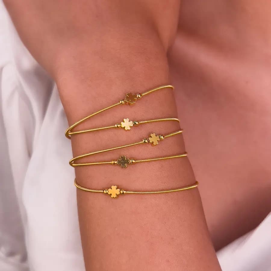 Pulseiras corda de violão douradas com trevo de quatro folhas, empilhadas no pulso de uma modelo